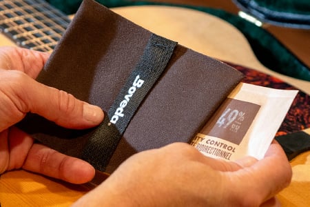 Male hand placing a 49% RH Boveda in a fabric holder with a guitar and guitar case behind his hands.