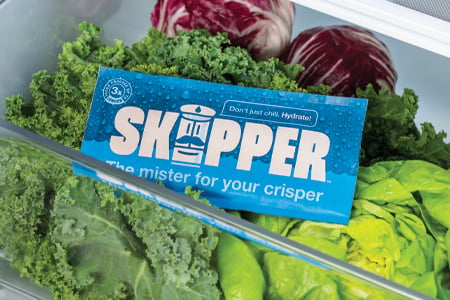 Skipper sitting on top of some vegetables in a fridge's crisper drawer